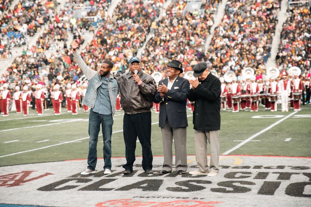 Four men stand on a football field during a ceremony, with a marching band and crowd visible in the background.