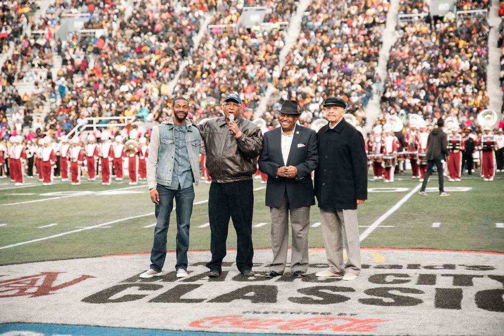 Four men pose on a football field at the CFL Classic game, with a marching band and stadium crowd in the background.