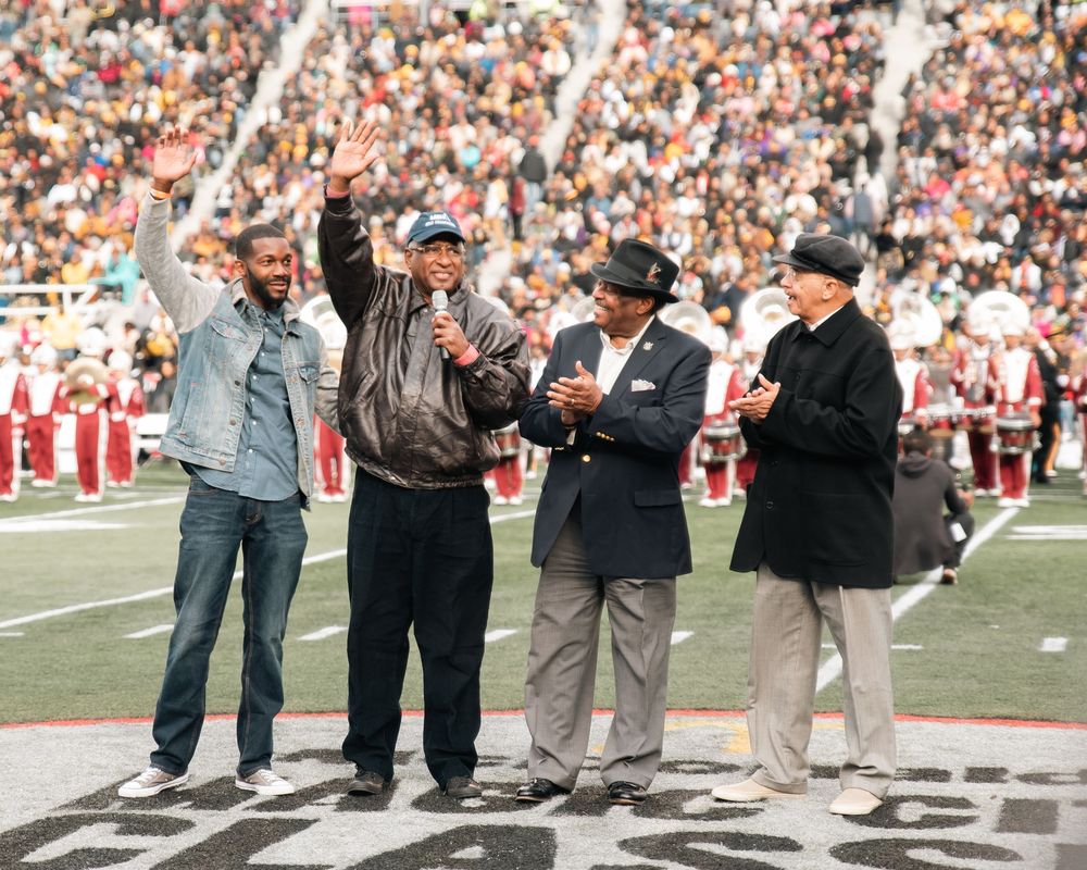Four men stand on a football field during a ceremony, one raising his hand while another holds a microphone.