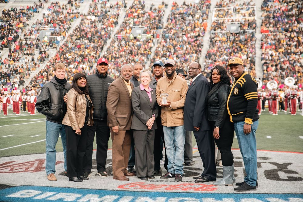 Group of people posing together on a football field during a game, with a large crowd visible in the background.