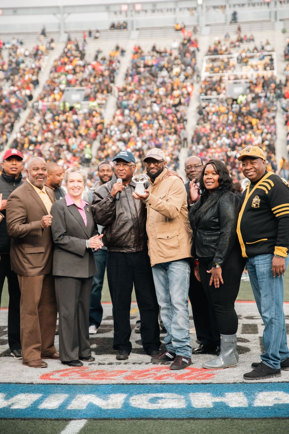 Group of people posing on football field during ceremony, packed stadium crowd visible in background.