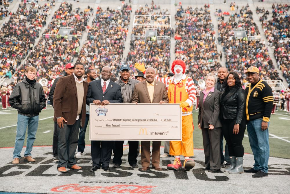 Group poses with Ronald McDonald and oversized charity check at football stadium fundraiser event.