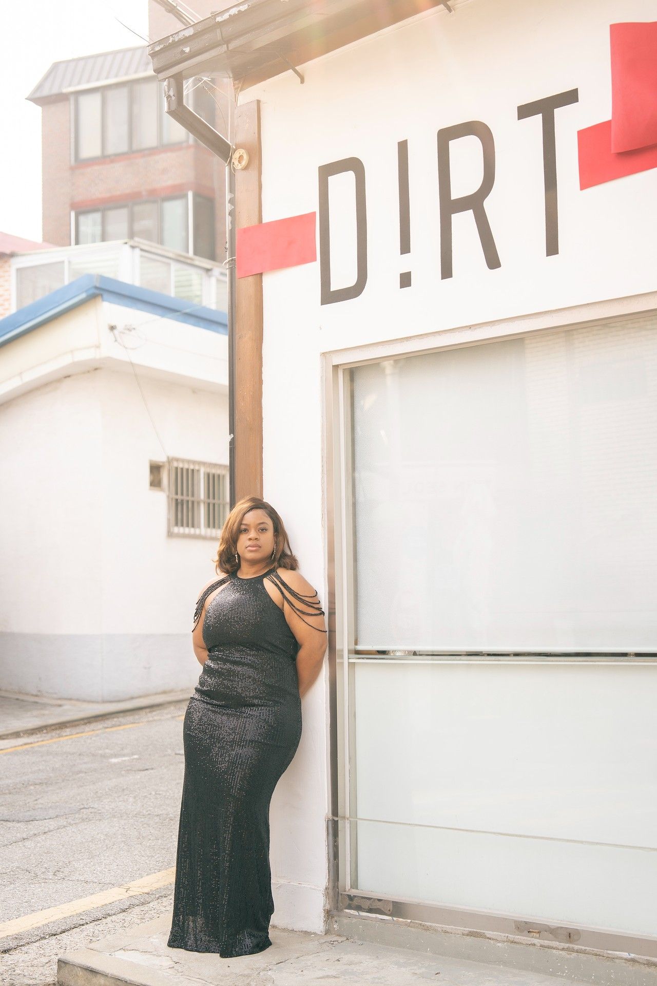 Woman in elegant black gown posing against white building with DIRT sign in urban outdoor setting.