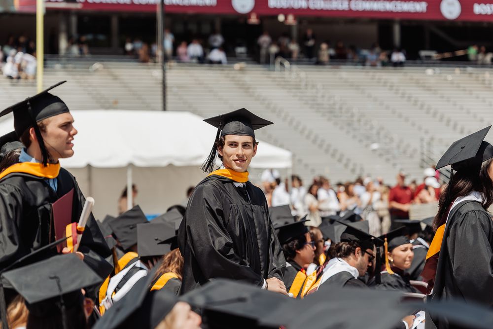 Students in caps and gowns at an outdoor college commencement ceremony on a sunny day.
