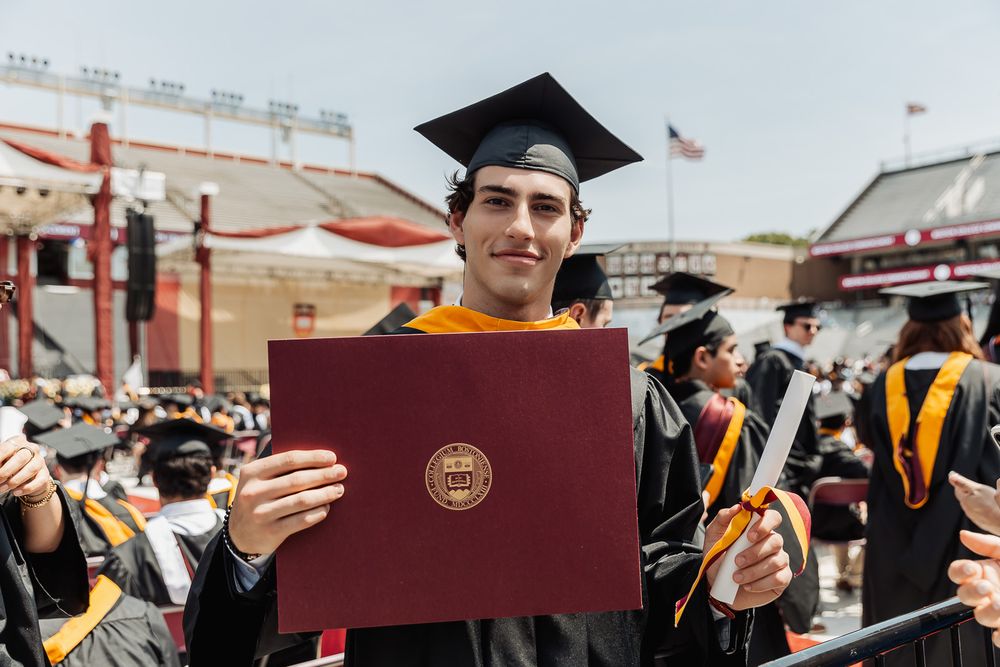 College graduate in cap and gown proudly holds diploma at outdoor commencement ceremony.