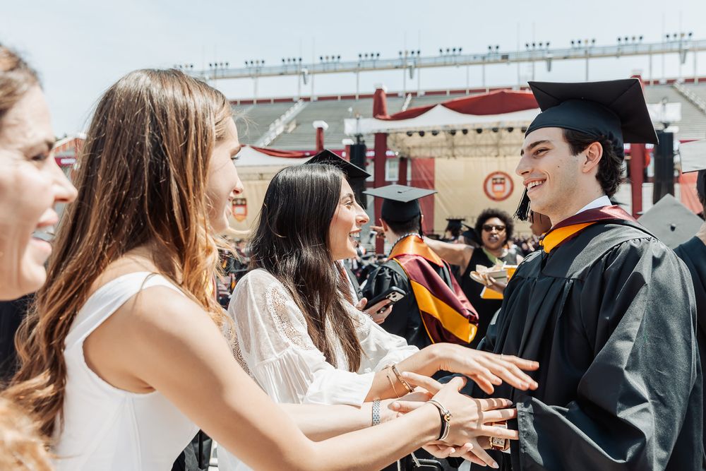 A smiling graduate in cap and gown shakes hands with guests at an outdoor graduation ceremony.