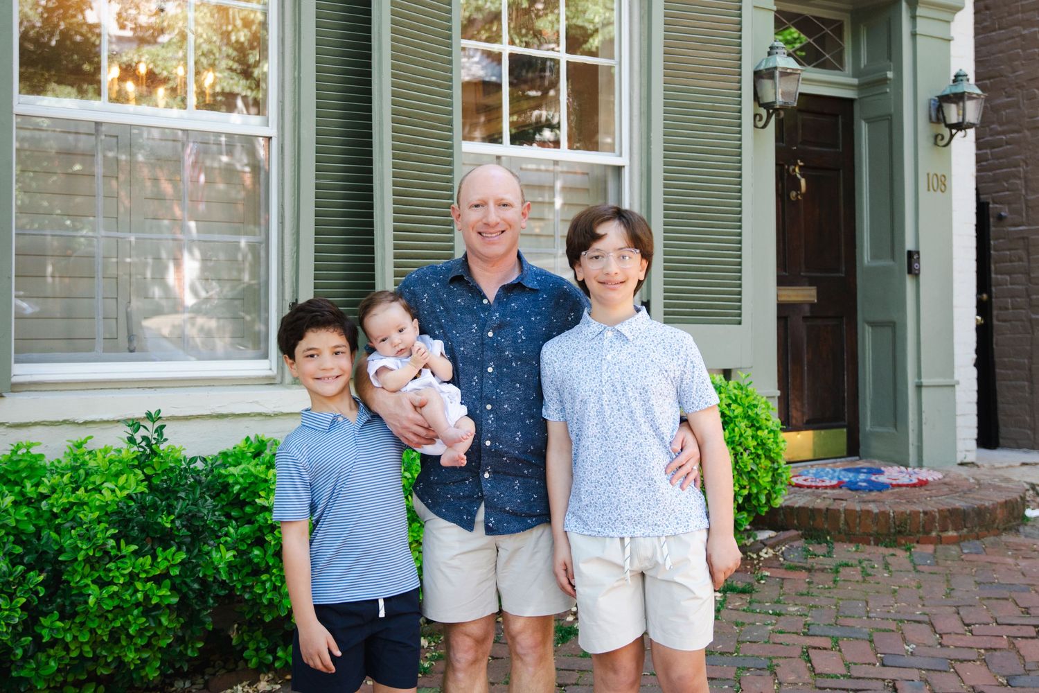 A smiling man poses with three children, including a baby, in front of a charming brick home with green shutters.