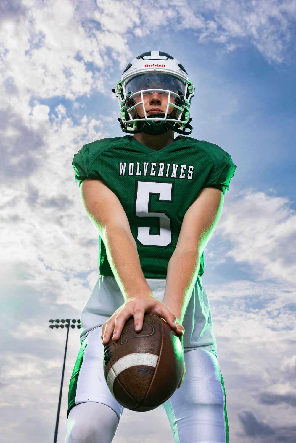 Wolverines #5 football player in green jersey and white helmet holds a football against a cloudy sky backdrop.
