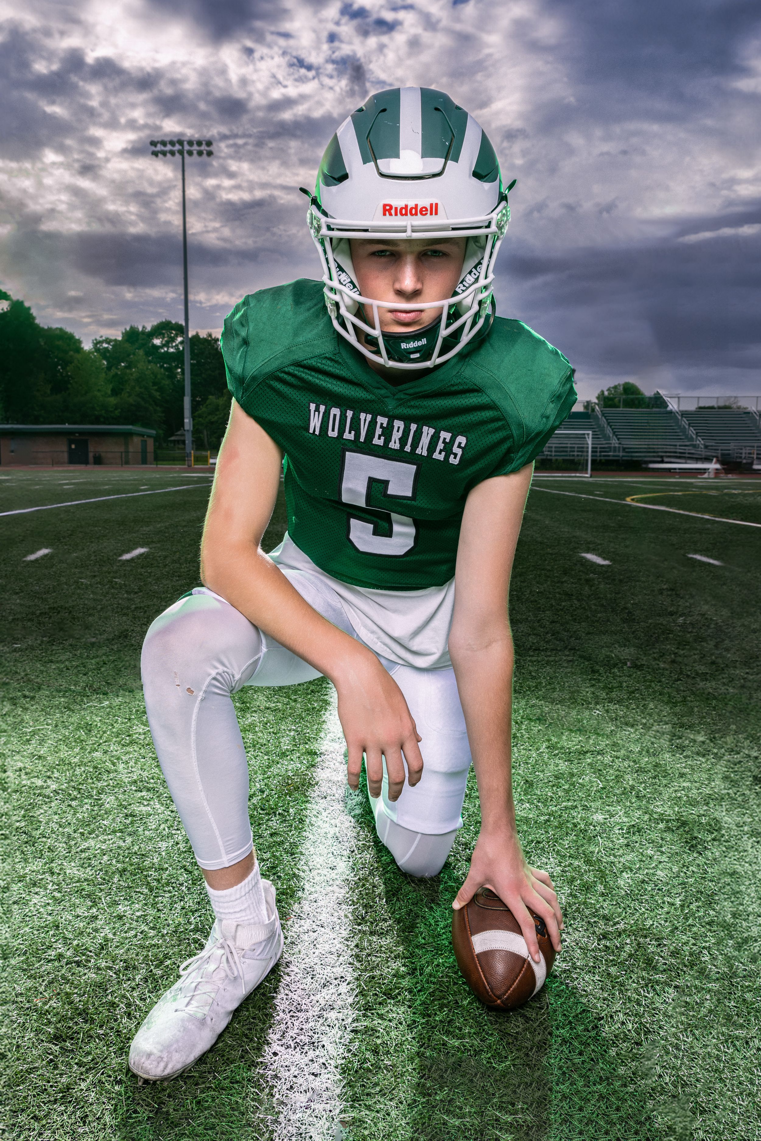 Young Wolverines football player #5 in green uniform kneeling on field holding ball wearing Riddell helmet.