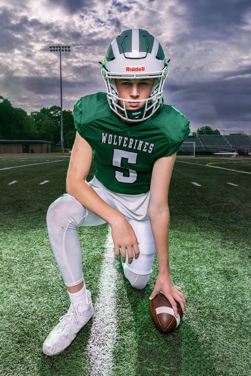 Young Wolverines football player #5 in green uniform kneeling on field holding ball wearing Riddell helmet.