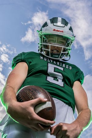 Wolverines #5 football player in green Riddell helmet holds a football, photographed from below against a cloudy sky.