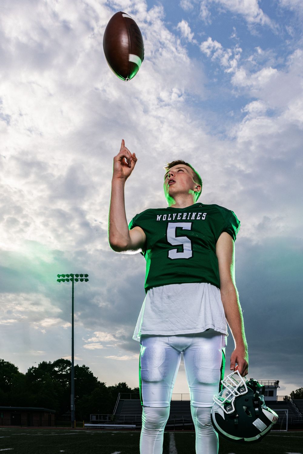 Young Wolverines football player #5 spinning a football on his finger while holding helmet under cloudy sky.