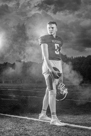 Black and white photo of a Wolverines football player #5 holding helmet on field with dramatic smoky sky backdrop.
