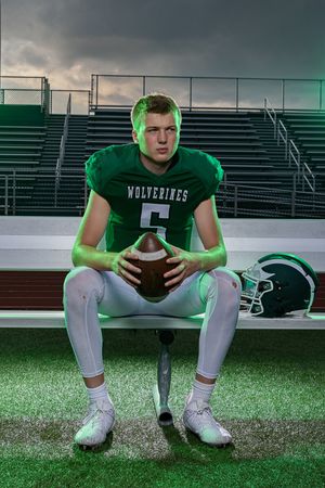 A Wolverines football player in green jersey number 5 sits on a bench holding a football at a stadium.
