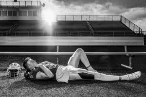 Black and white photo of a football player resting on a field, helmet beside him, with stadium bleachers in the background.
