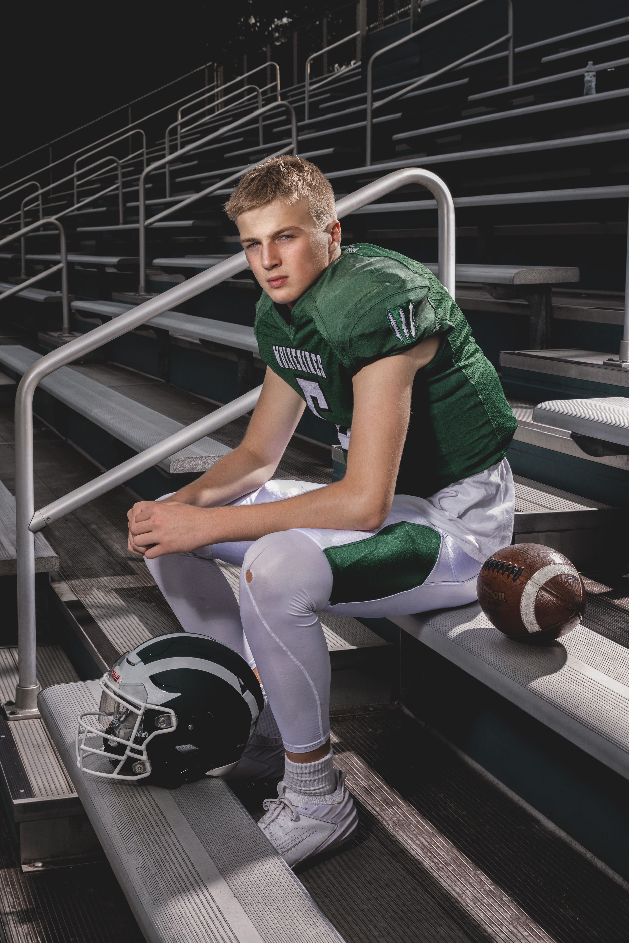 Young football player in green jersey sits on bleachers with helmet and ball, posing for a portrait photo.