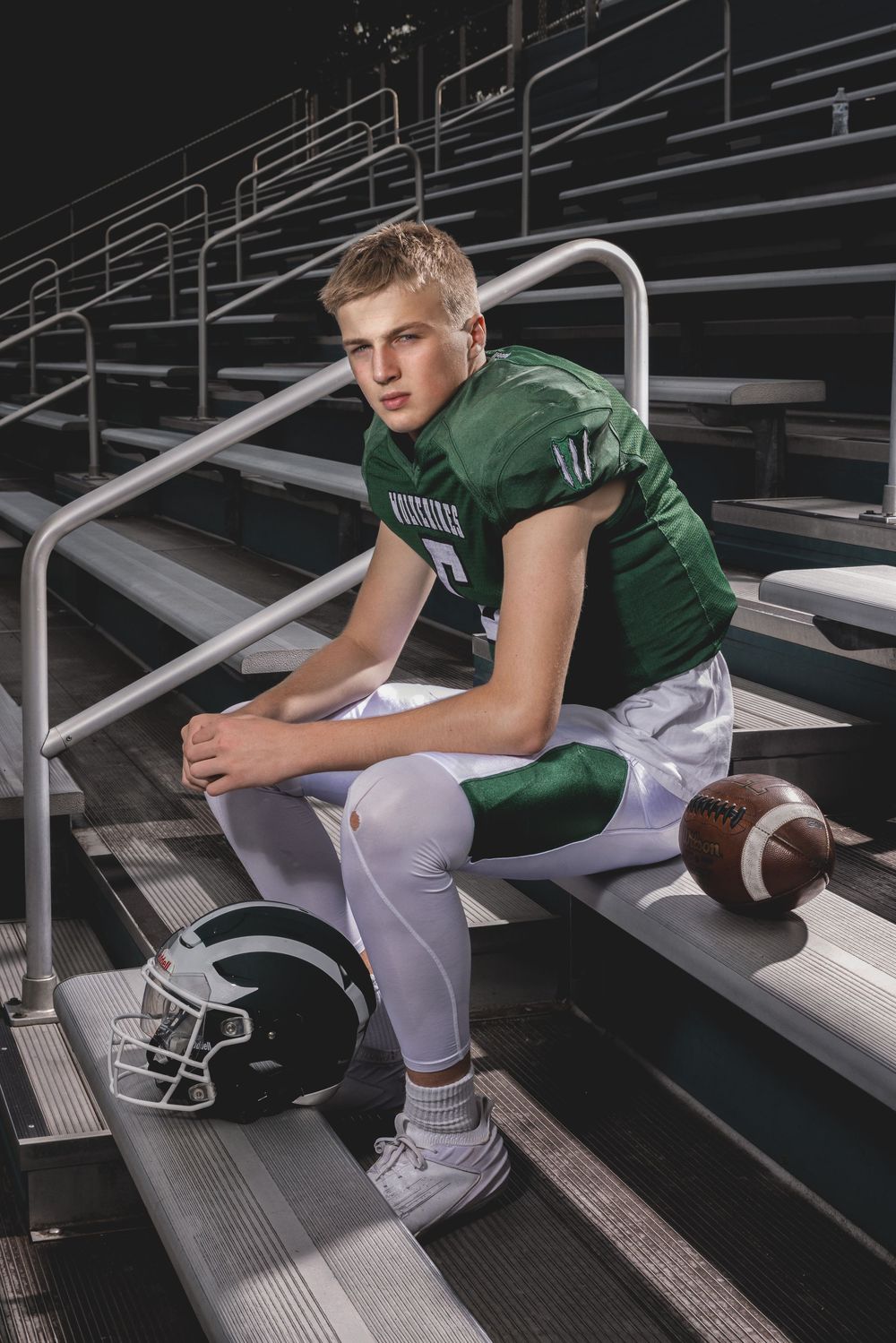 Young football player in green jersey sits on bleachers with helmet and ball, posing for a portrait photo.