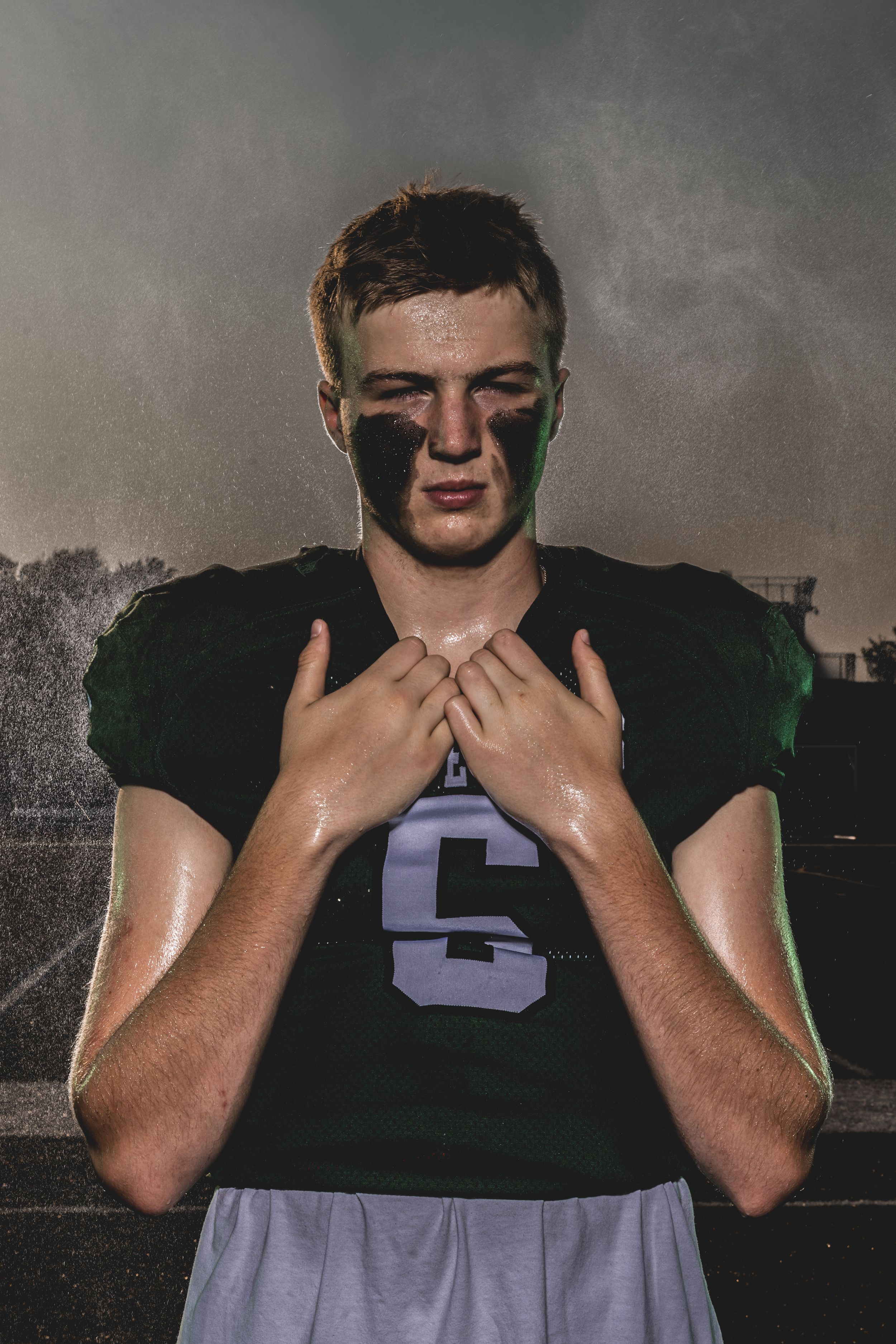 A young football player in a green jersey with eye black, hands clasped at chest, standing under dramatic moody lighting.