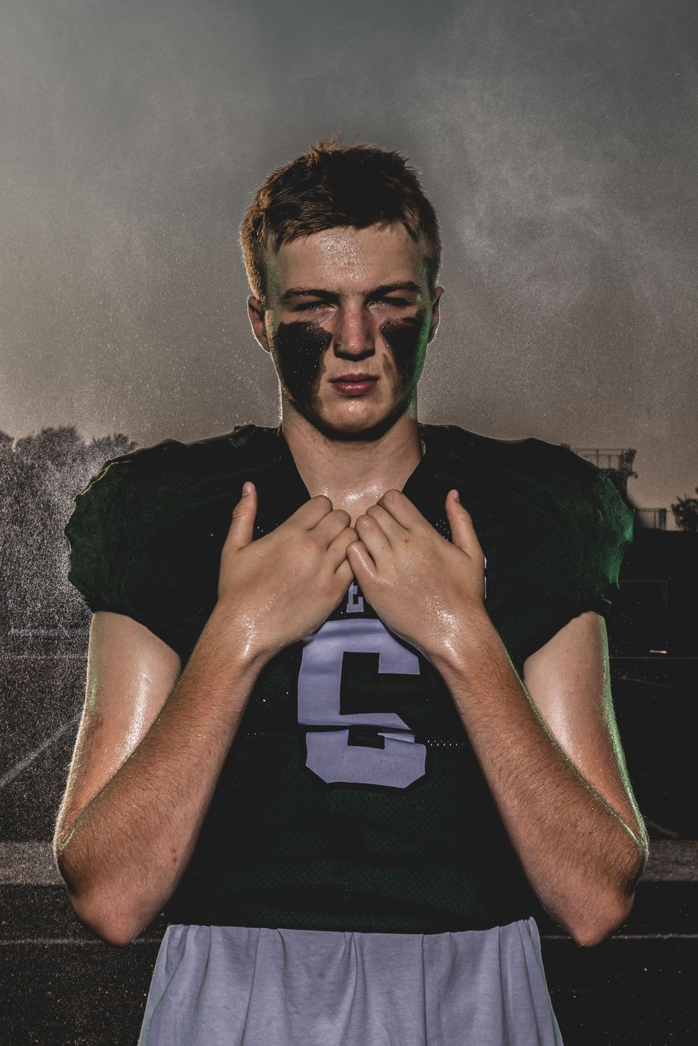 A young football player in a green jersey with eye black, hands clasped at chest, standing under dramatic moody lighting.
