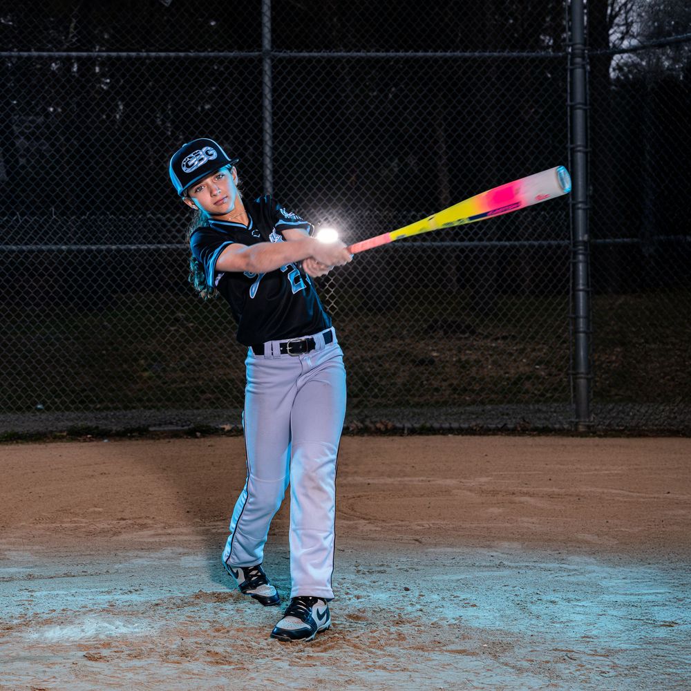 Young baseball player swinging a glowing light-up bat at night on a baseball field wearing helmet and uniform.