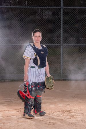 Female softball catcher in full gear stands on dirt field holding helmet and mitt, wearing pinstripe uniform.