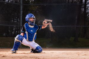 Baseball catcher in blue gear crouching to catch a pitch on a dirt field, with dust flying from the impact.