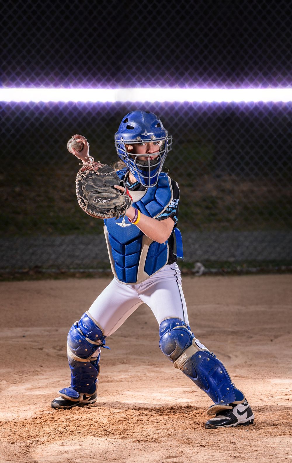 Youth baseball catcher in blue gear crouches in ready position on dirt field under bright stadium lights at night.