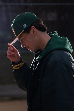 Young male baseball player in green and gold cap and jacket looking down on dark background.