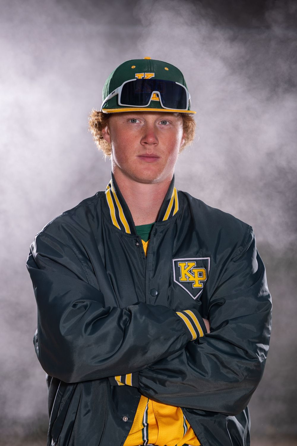 Young male baseball player in green and gold KP jacket and cap poses with arms crossed against smoky background.