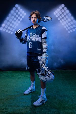 Young lacrosse player in blue jersey number 6 holding stick and helmet under dramatic stadium lights.