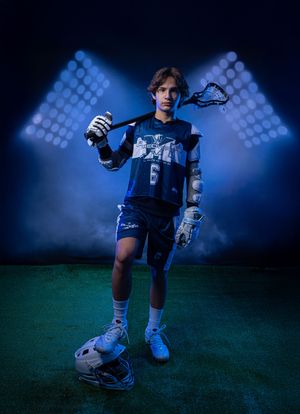 Young lacrosse player in blue uniform holding stick under dramatic stadium lights with misty background.