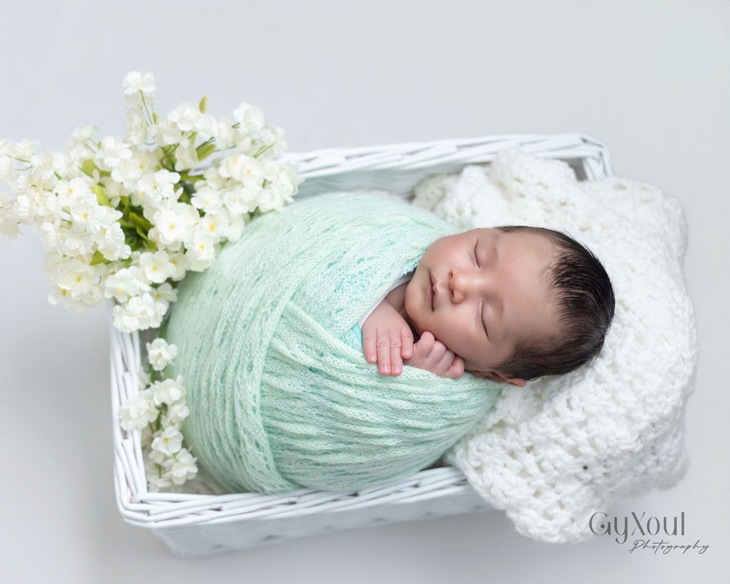 Sleeping newborn baby swaddled in mint green wrap resting in white wicker basket with white flowers.