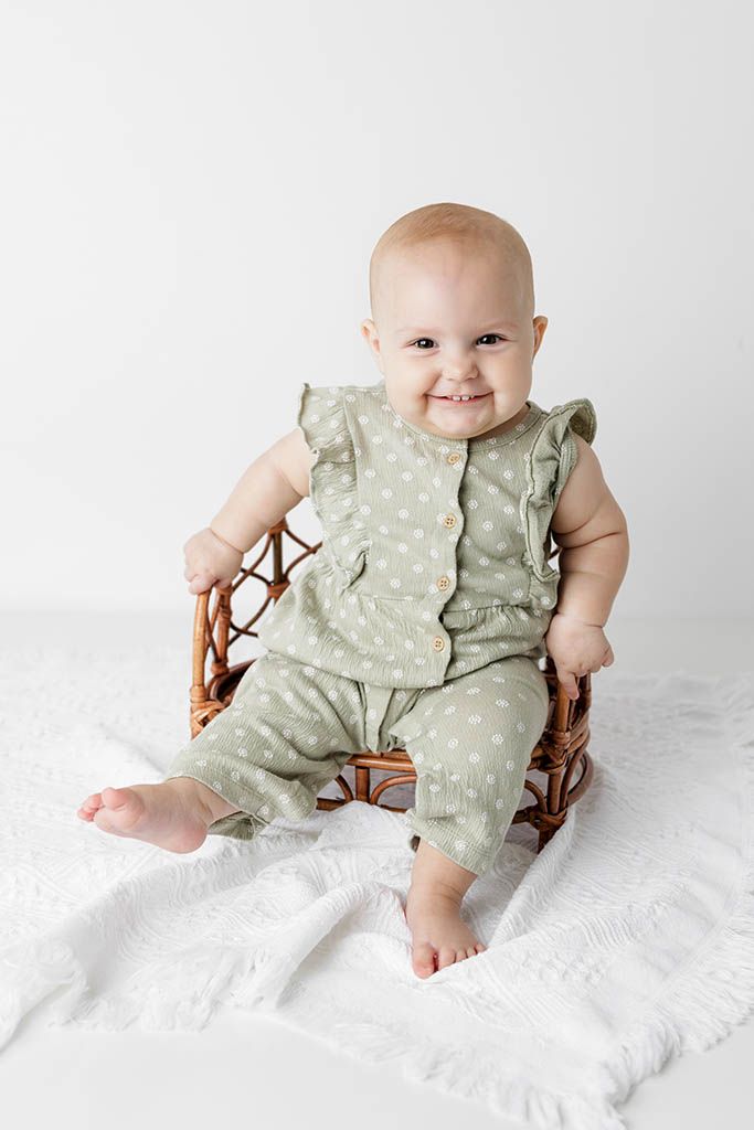Happy baby in sage green polka dot romper sitting in a wicker chair on white blanket.