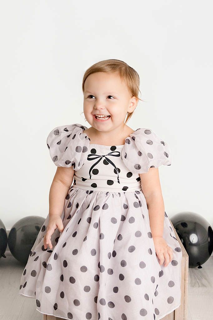 Happy toddler girl in polka dot dress laughing surrounded by black balloons by Temecula child photographer.