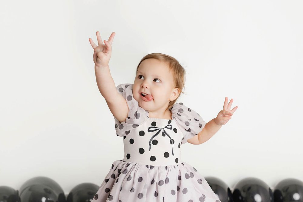 Joyful toddler girl in polka dot dress raises hands surrounded by black balloons against white background.