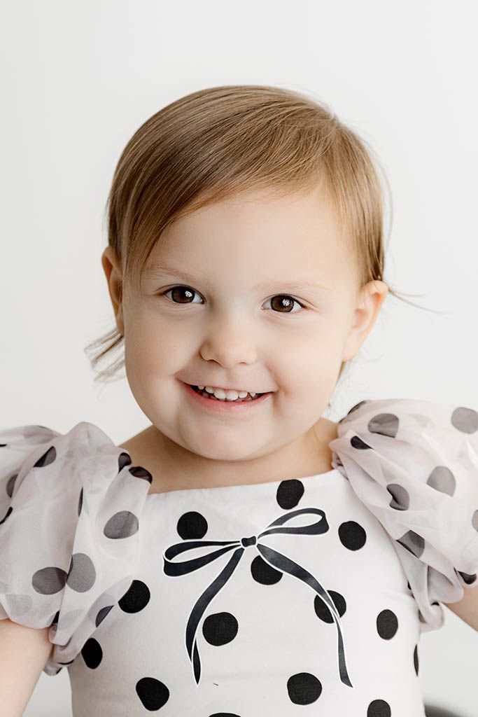 Cute toddler girl smiling in a white and black polka dot dress with bow detail.