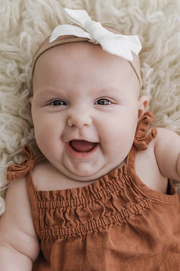Smiling baby girl in rust romper and white bow headband lying on fluffy cream blanket.