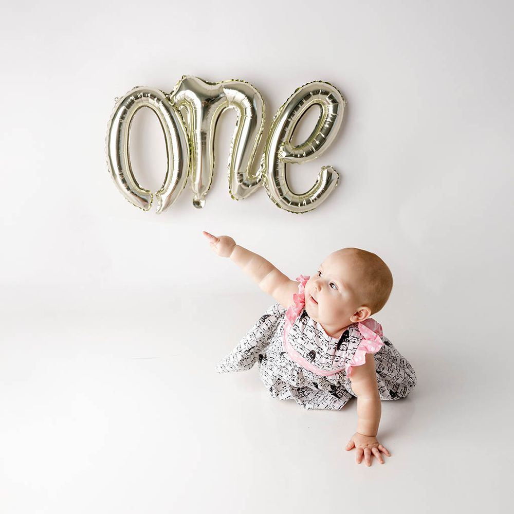 Baby girl in floral dress pointing at a gold 'one' balloon during her first birthday photo shoot by Temecula child photographer.