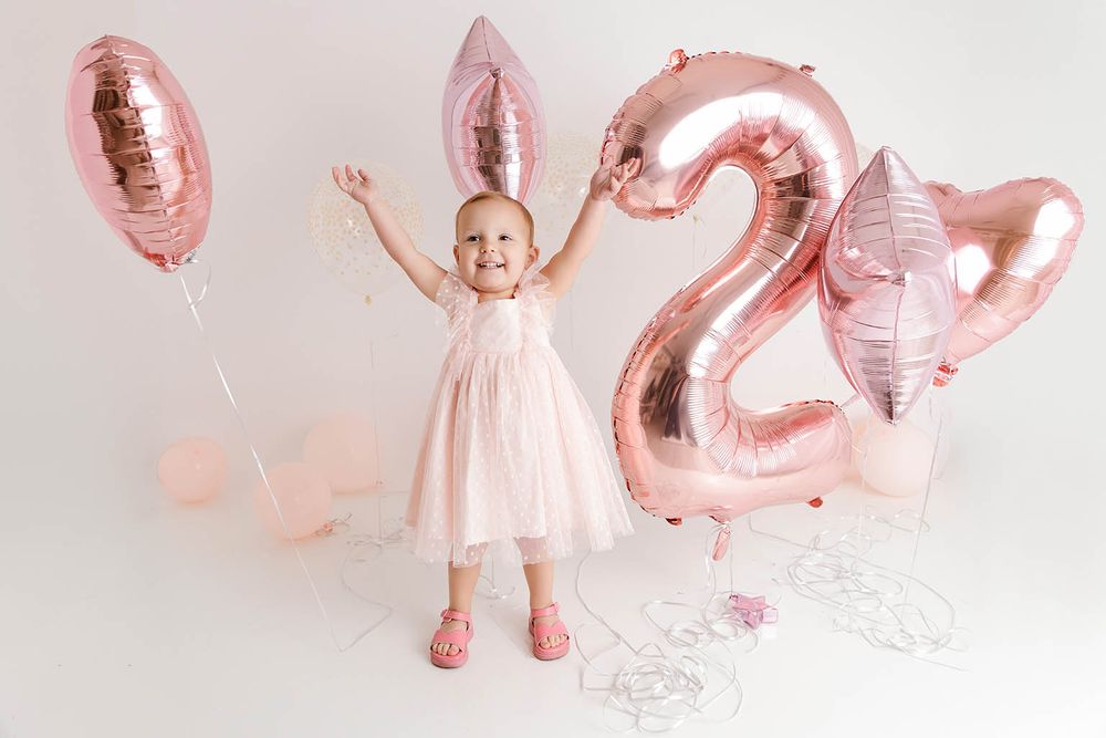 Toddler girl in pink dress with bunny ears celebrates 2nd birthday with rose gold number balloons.