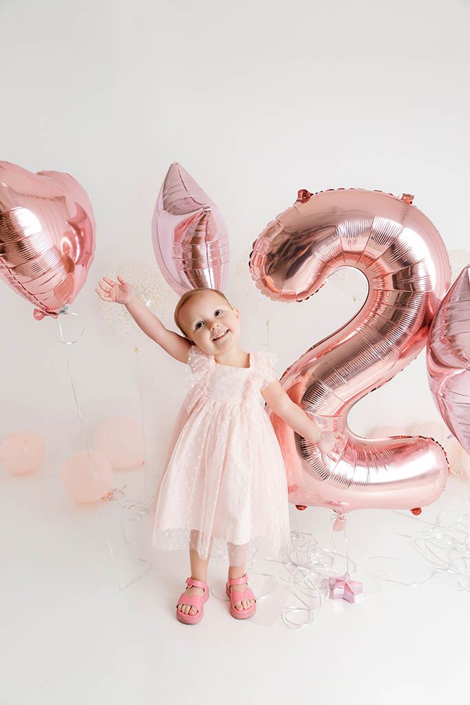 Smiling toddler girl in pink dress celebrating 2nd birthday with rose gold number balloon.