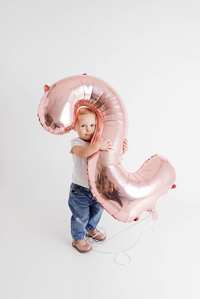 Toddler child hugging a large rose gold number 2 balloon on white background.