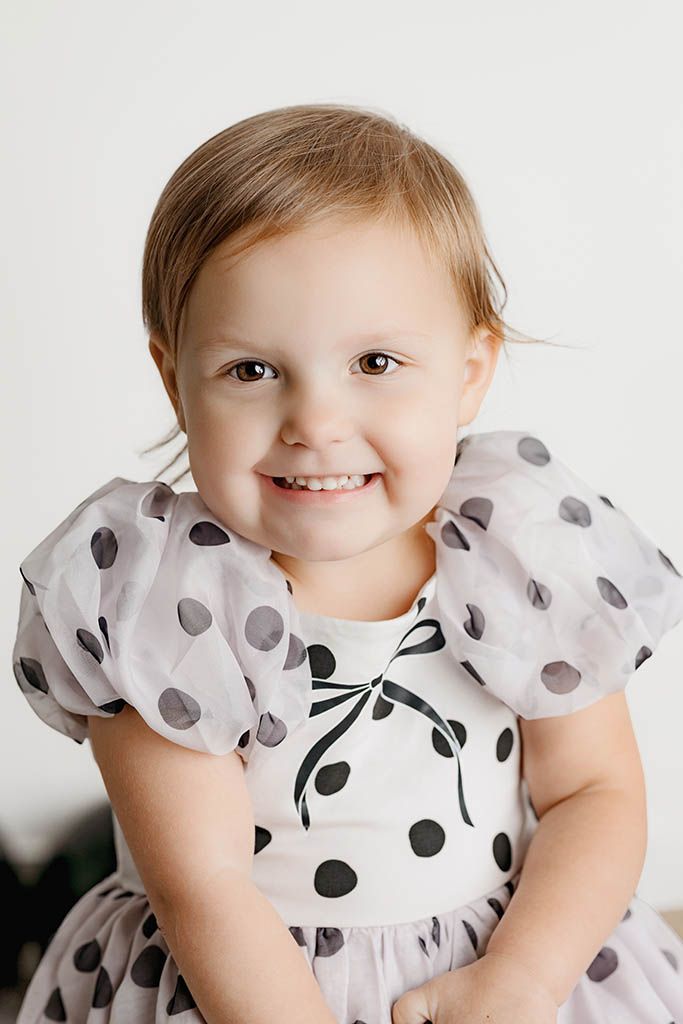 Cute toddler girl smiling in a white and black polka dot dress with bow during portrait session.