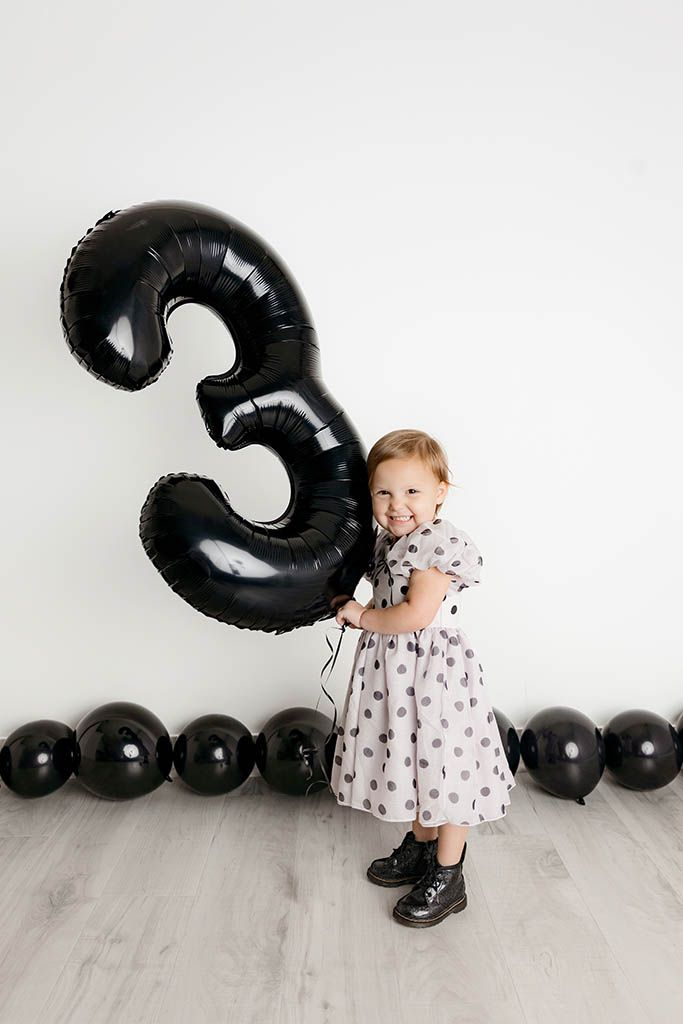 Smiling toddler girl in polka dot dress holding large black number 3 balloon by Temecula child photographer.