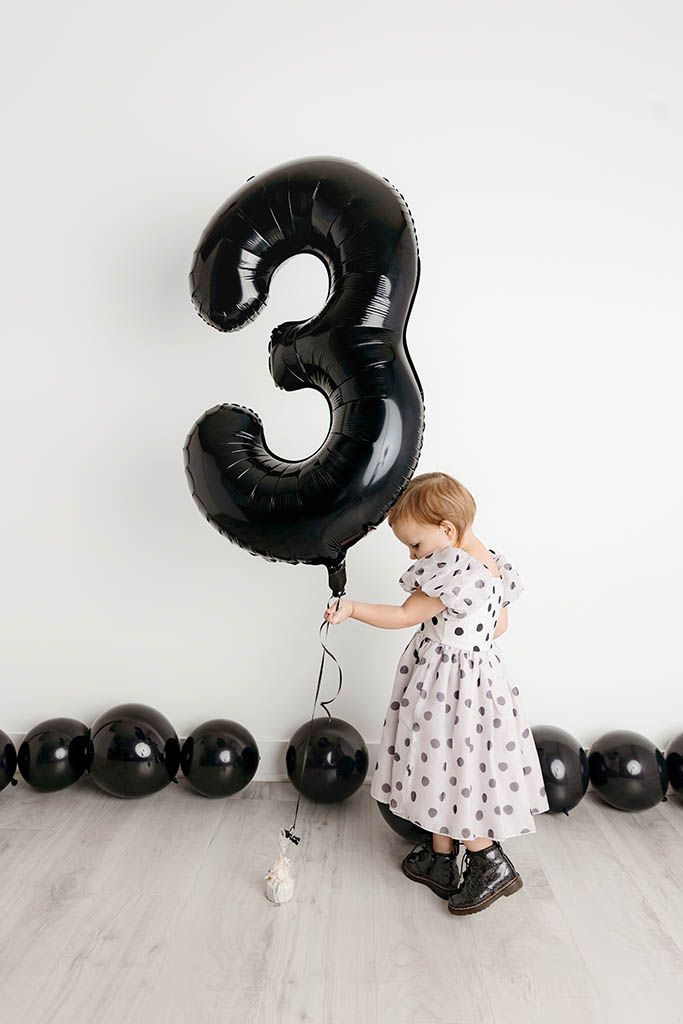 Toddler girl in polka dot dress bending down near large black number 3 balloon by Temecula child photographer.