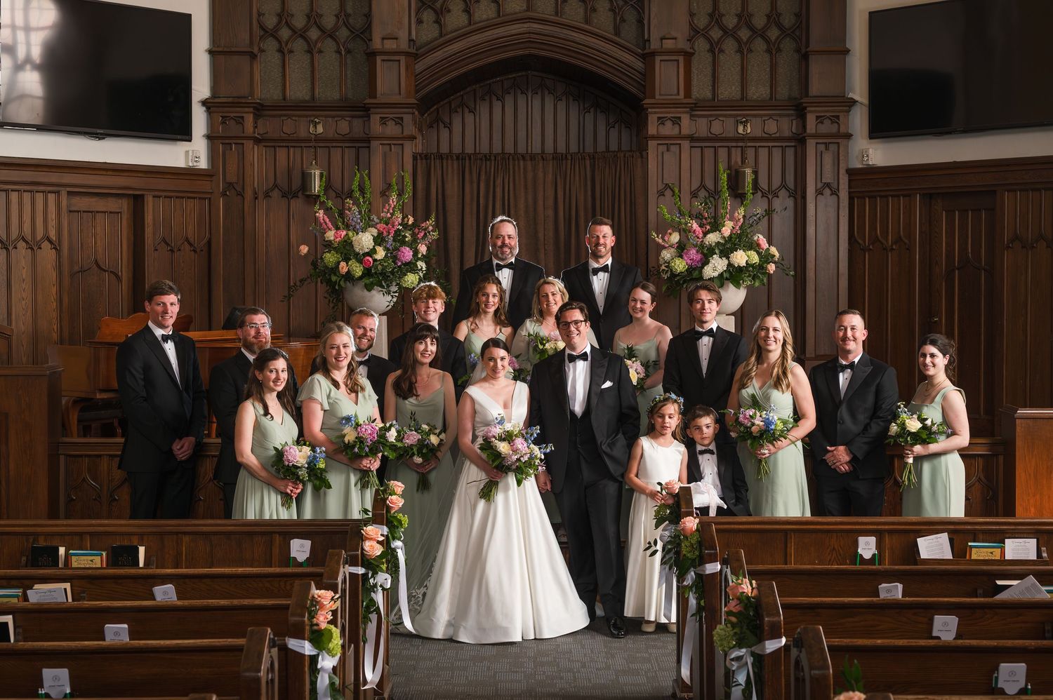 Wedding party poses at church altar with floral arrangements, bride in white gown, groom in tuxedo, surrounded by family.