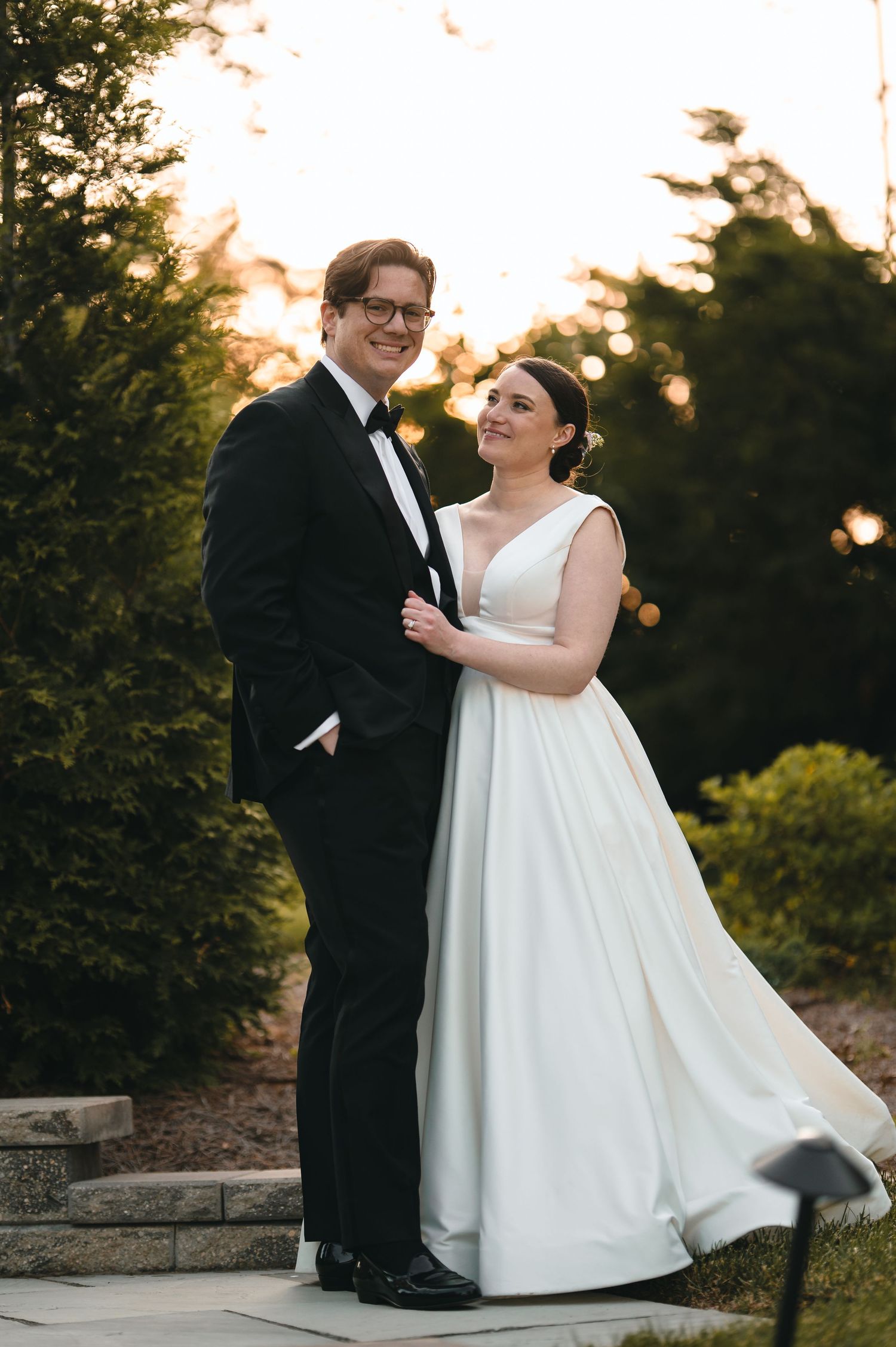 Bride and groom pose together outdoors at sunset, she in a white ballgown, he in a classic black tuxedo.