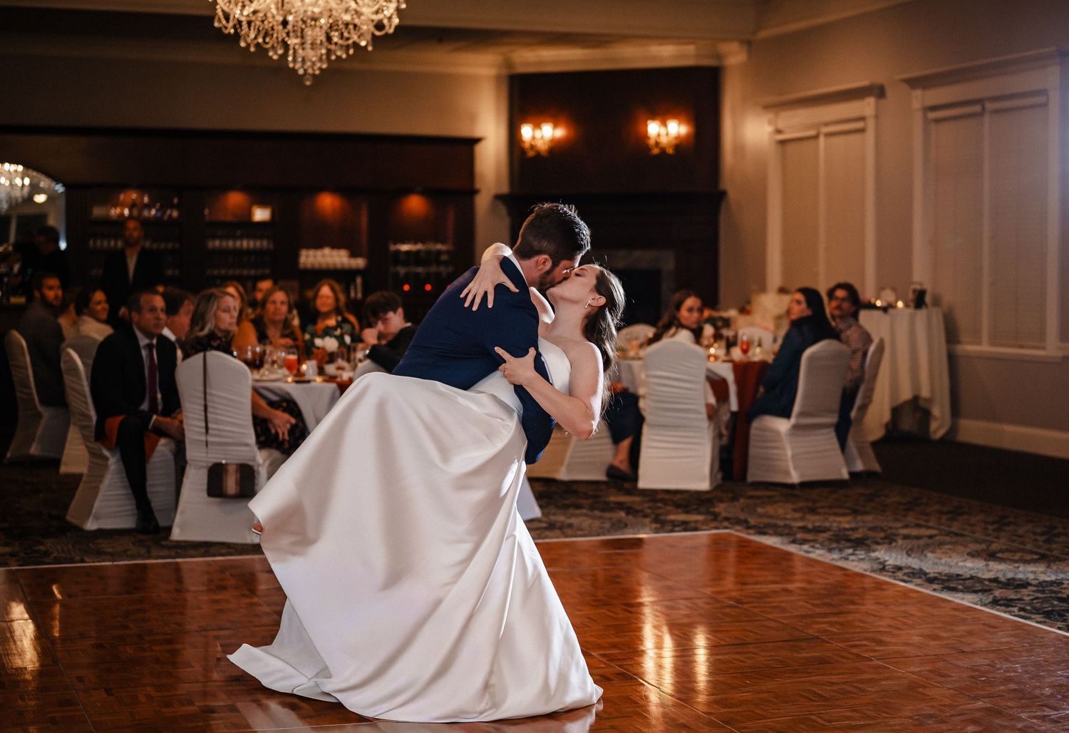Bride and groom share romantic first dance dip kiss in elegant ballroom wedding reception with guests seated nearby.