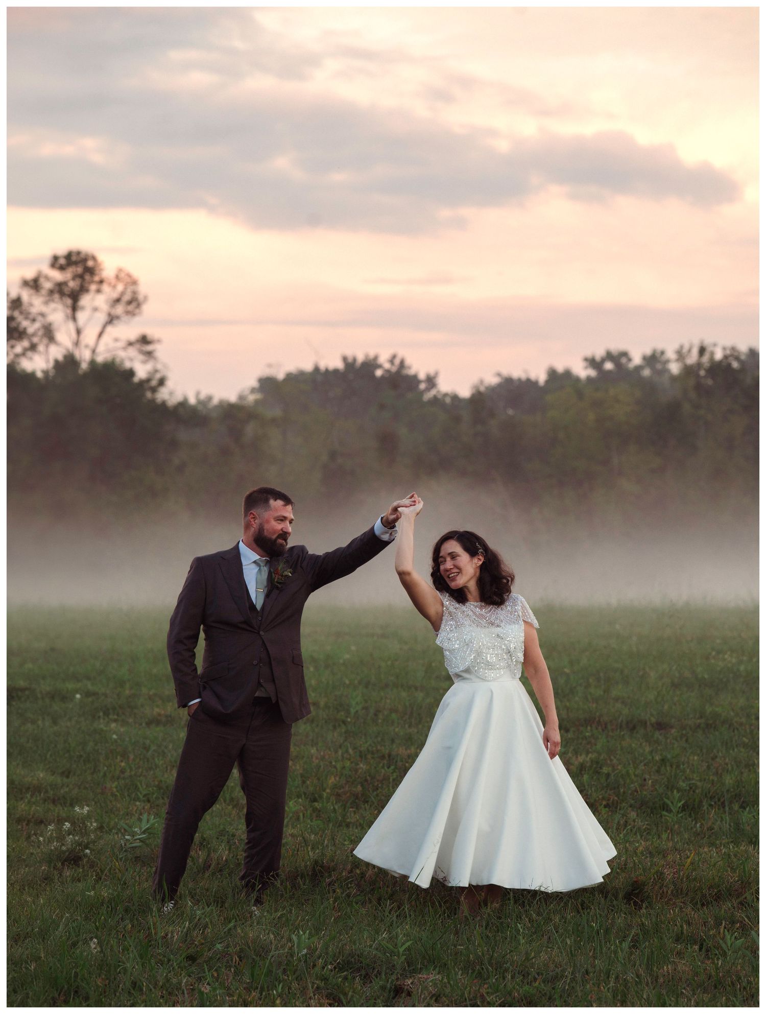 A couple dances joyfully in a misty field at sunset, the bride in a tea-length white dress and groom in a dark suit.