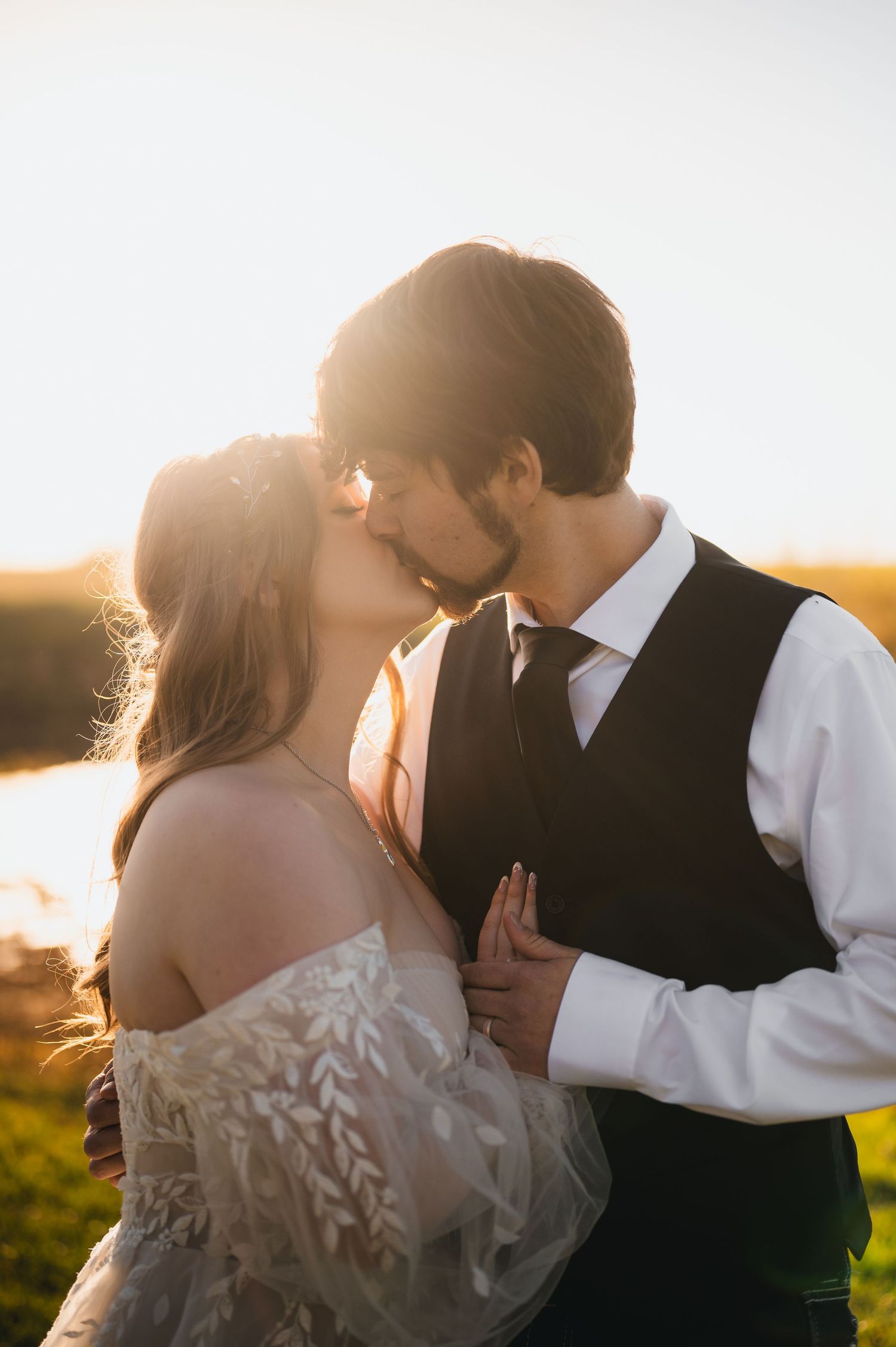 Bride and groom sharing a romantic sunset kiss outdoors, bride in lace gown, groom in white shirt and dark vest.