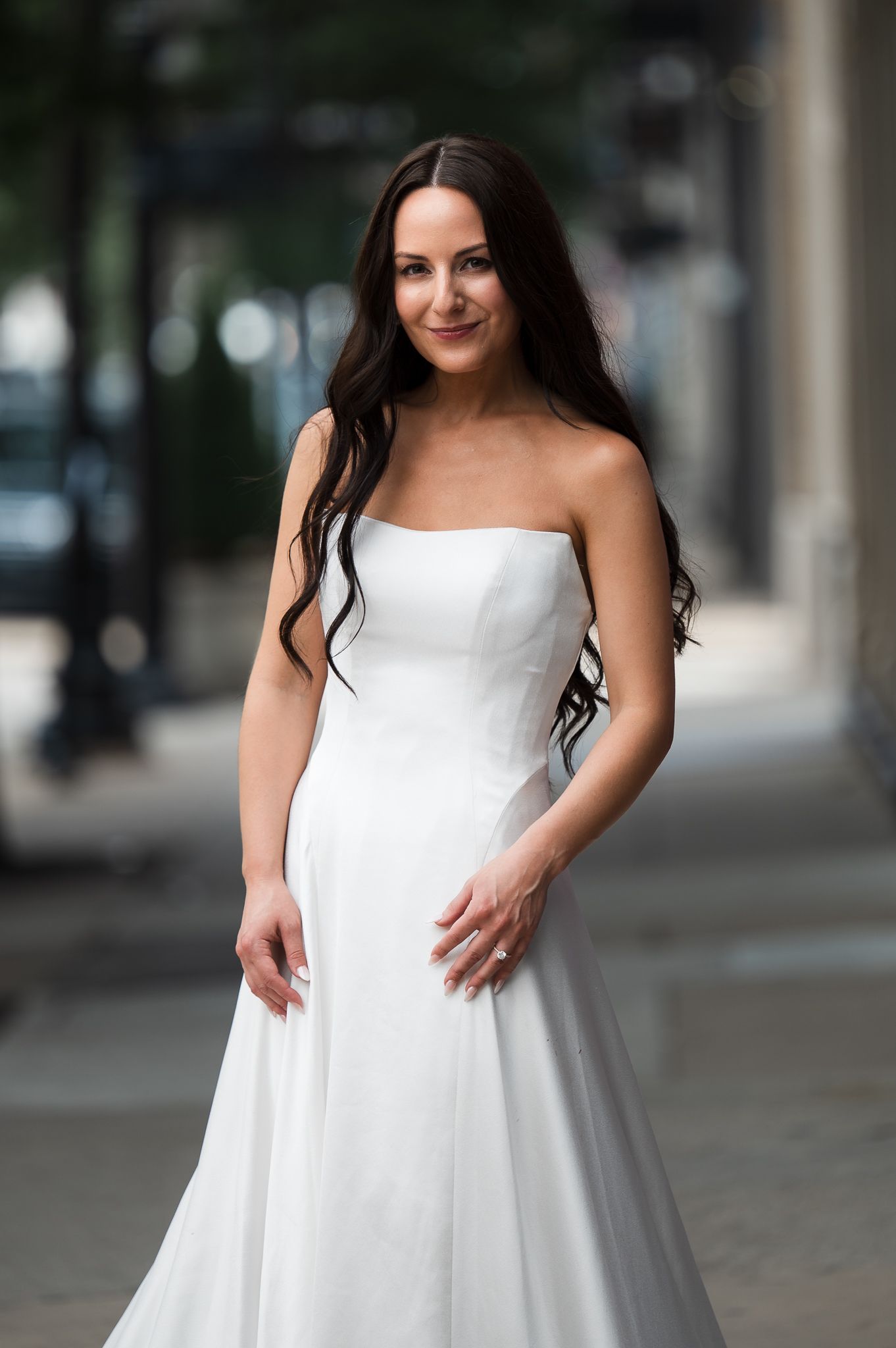 Smiling bride in strapless white A-line wedding gown posing outdoors on an urban street.